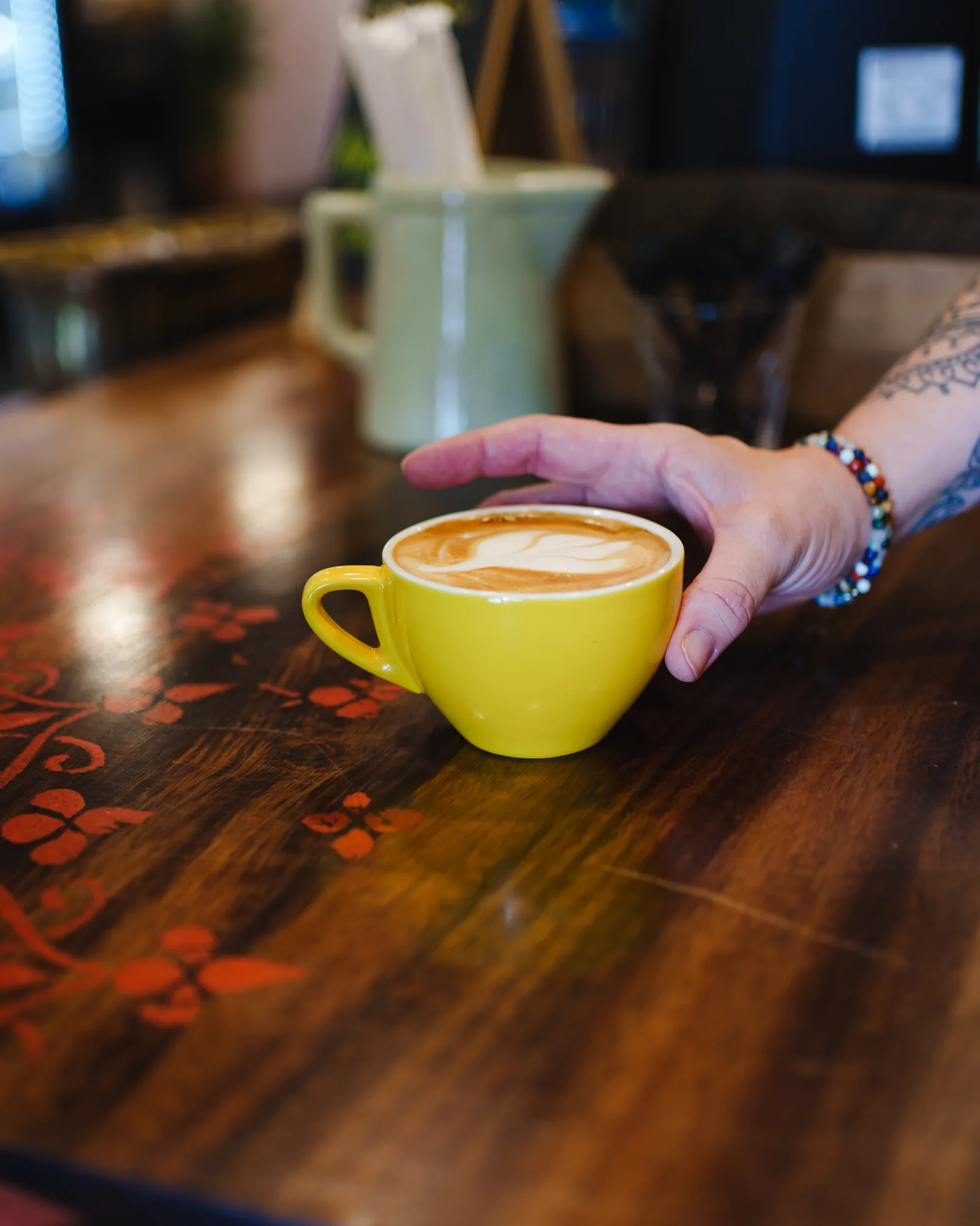A latte in a yellow cup being set down on a stencilled wood table